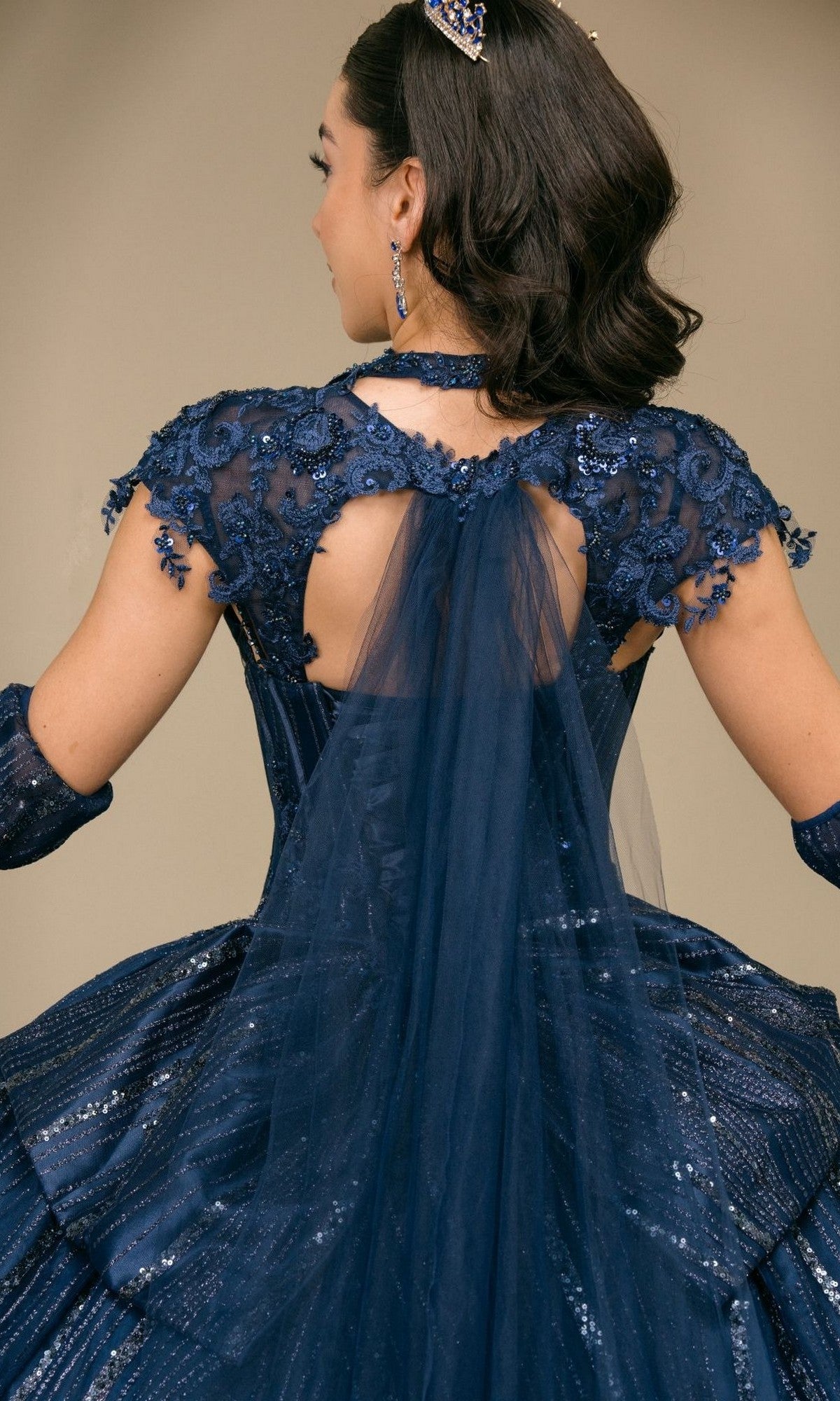 Wearing the Quinceanera Dress 8510JQ By Cinderella Couture, a woman is shown from behind in a navy blue sequined gown featuring lace and tulle details, accessorized with matching pieces and a jeweled headband for the perfect quinceanera ball gown look.